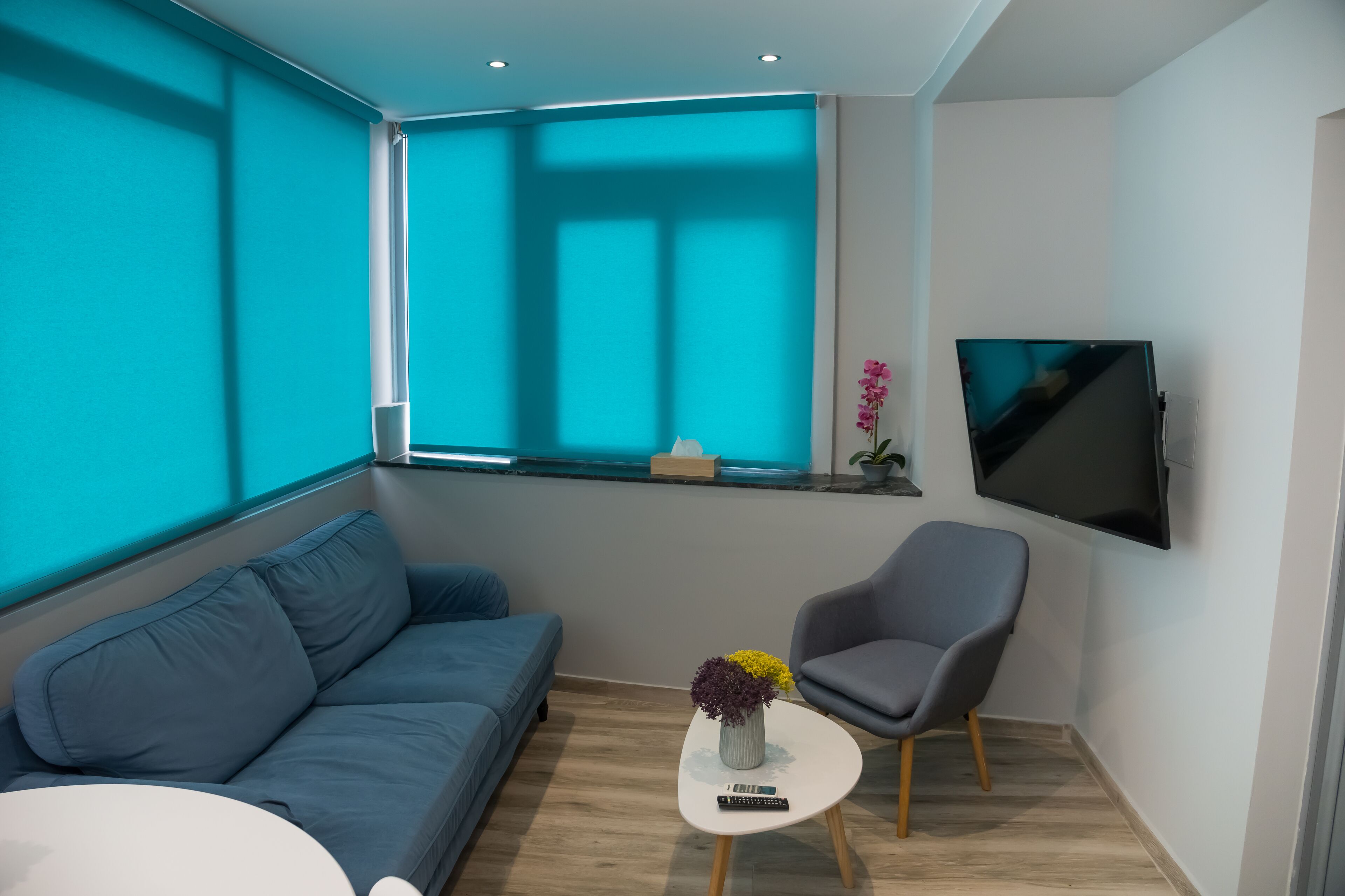 Un saló modern amb un sofà blau, un armari gris, una taula petita blanca amb un ram de flors, un televisor penjat i una finestra amb vista al cel.
