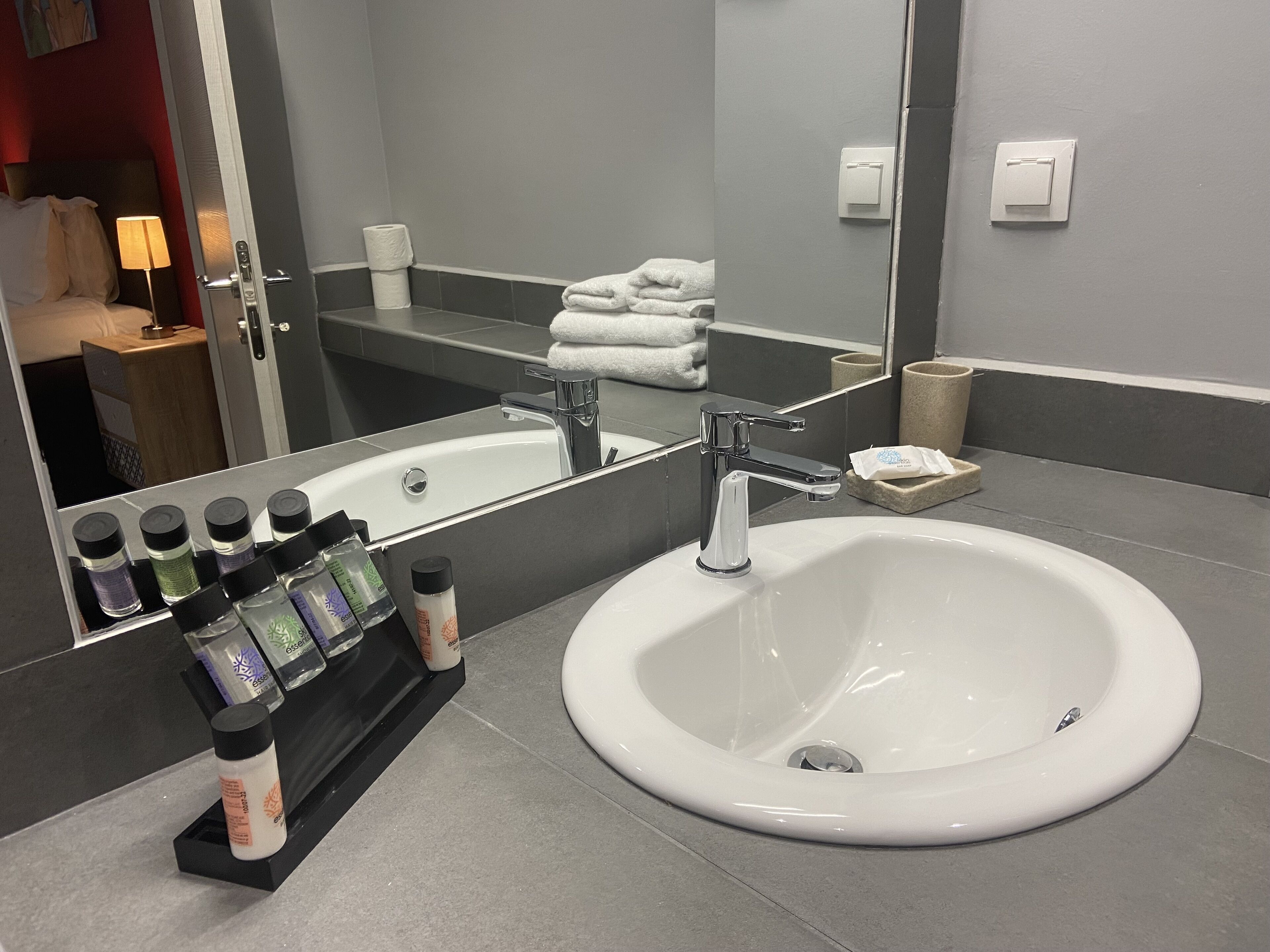 A modern bathroom with a large white sink, chrome faucet, and a mirror reflecting the room. There are toiletries on the counter and a towel rack with folded towels.