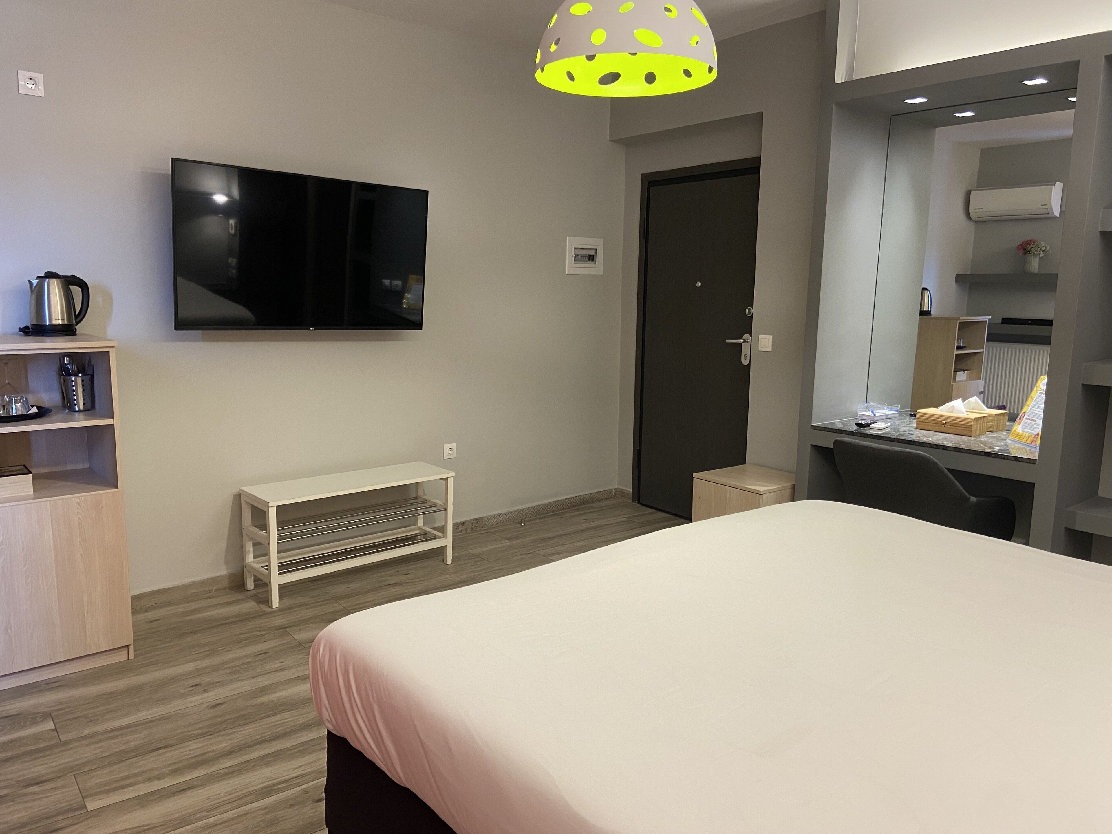 A modern hotel room with a large bed, a flat-screen TV mounted on the wall, a small bedside table, a wooden nightstand, and a built-in desk with a chair.