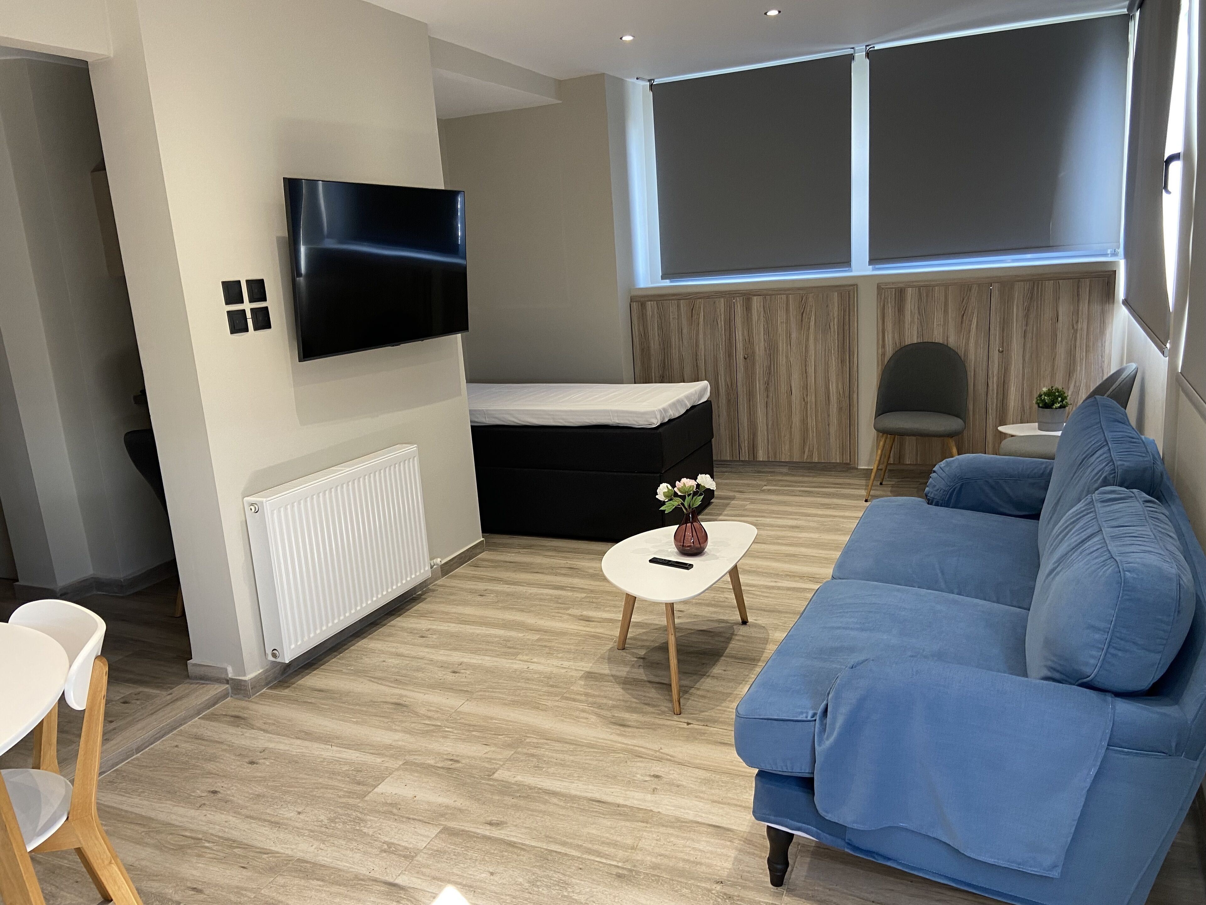 A modern living room with a blue sofa, a black ottoman, a small white table with a vase, and a TV mounted on the wall.