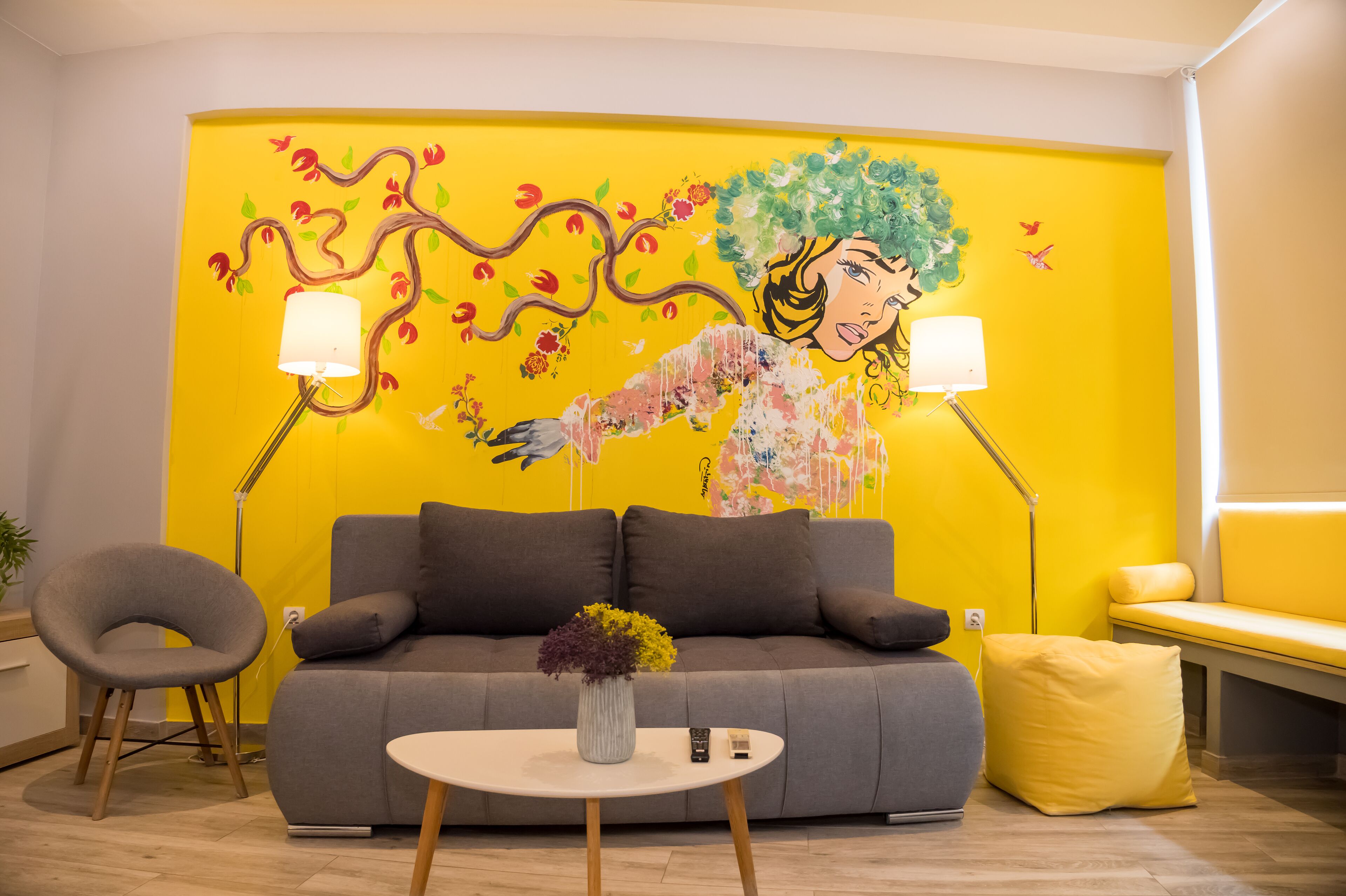 A modern living room with a vibrant yellow wall, a grey sofa, a round white coffee table, and a yellow pouf.