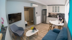 A modern living room with a sofa, a coffee table, and a TV. There is a kitchen area with cabinets and a refrigerator. - Connect Studios & Apartments (Athens)