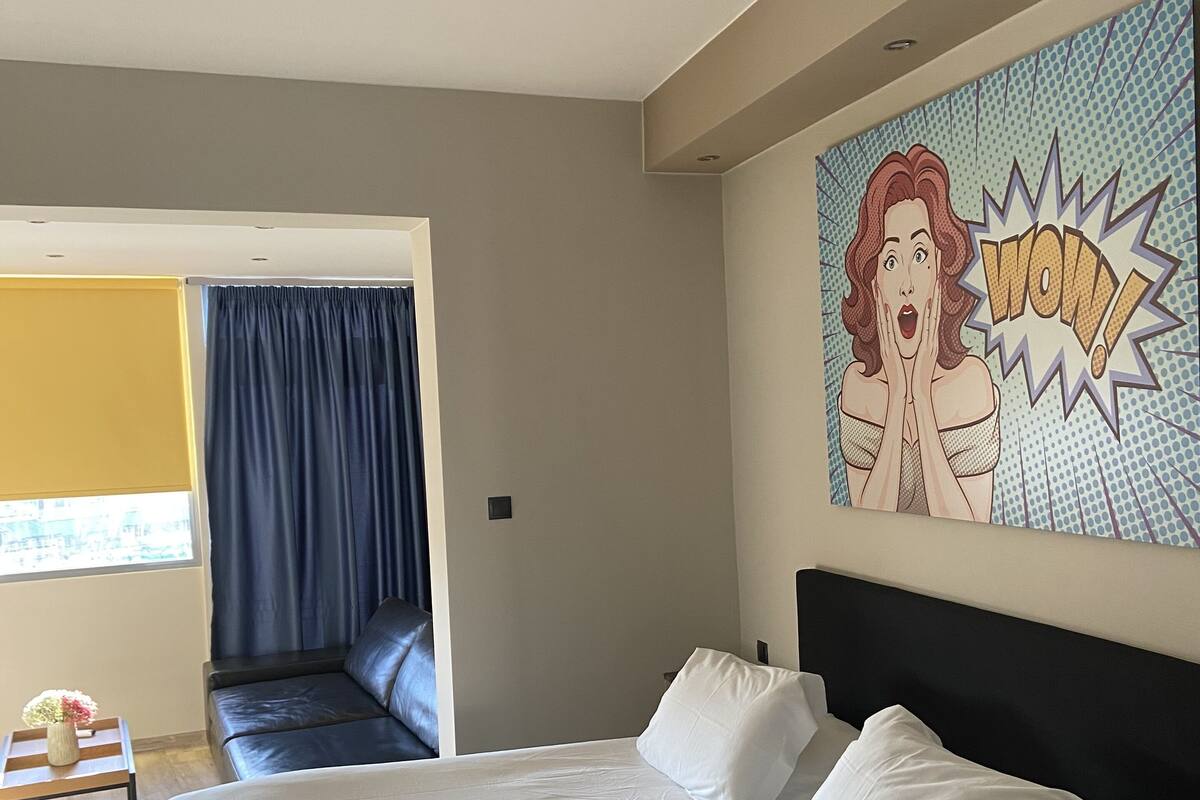 Habitación de hotel con una cama grande, un cuadro en la pared, una ventana con cortinas y una pequeña mesa con un florero.