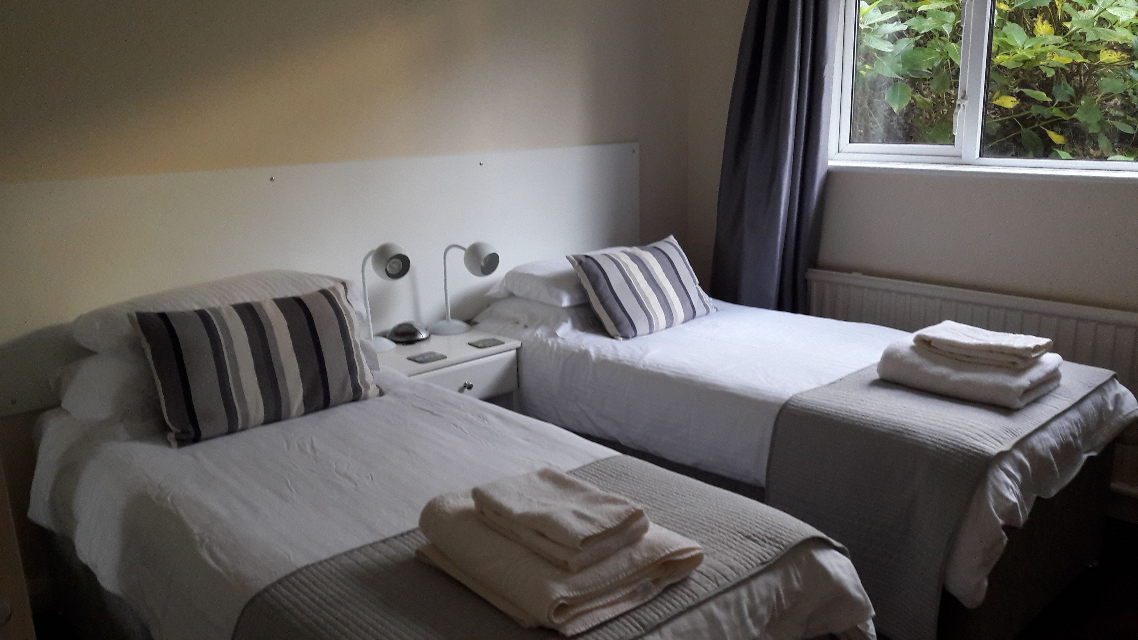 Habitación de hotel con dos camas individuales, una mesita de noche con lámpara, una ventana con cortinas y un radiador.