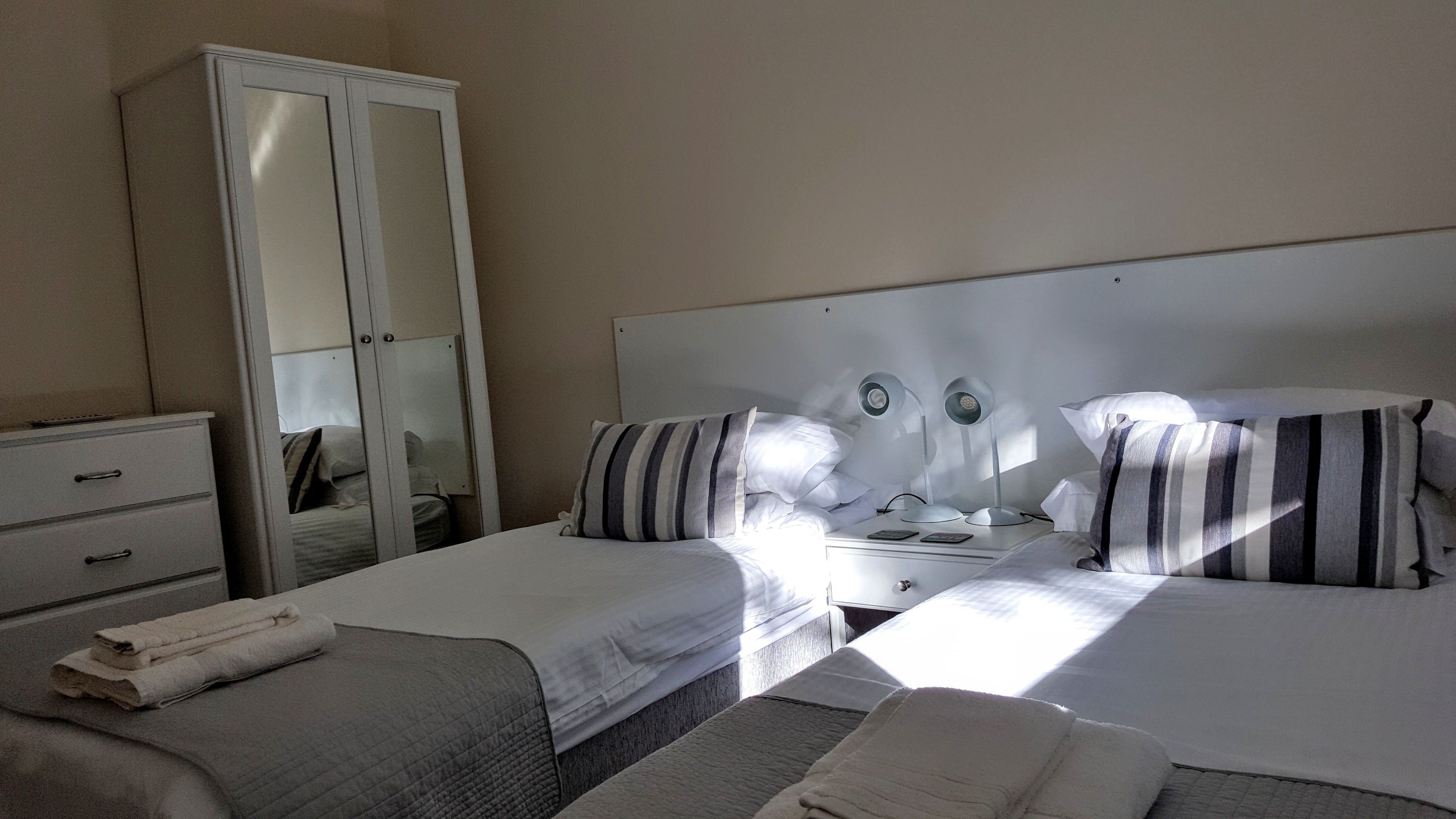 A hotel room with two beds, a wardrobe, a bedside table with lamps, and a mirror.