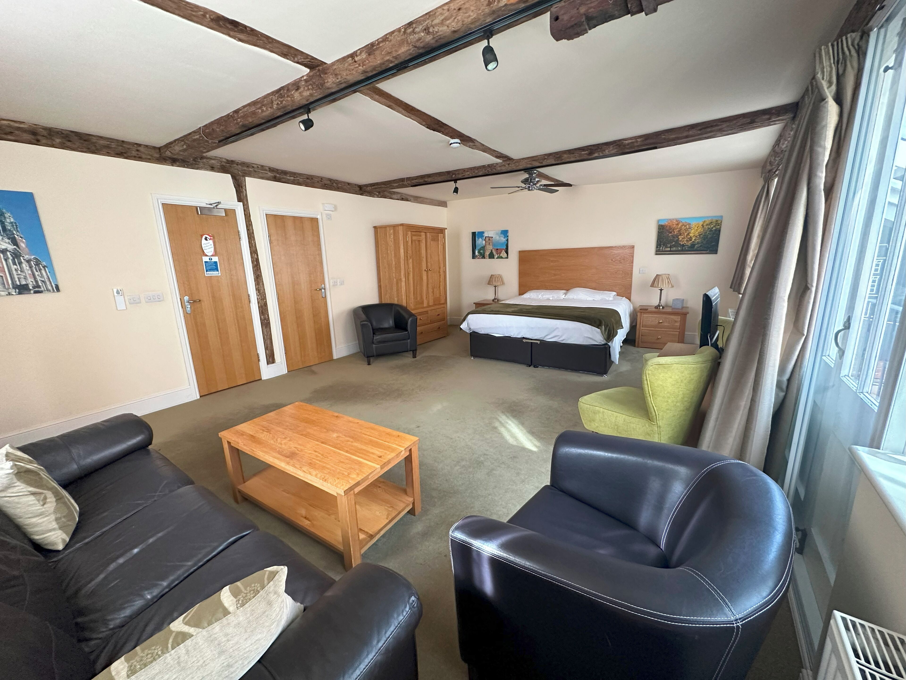 A room with a bed, a wooden table, two sofas, a green armchair, and a ceiling fan.