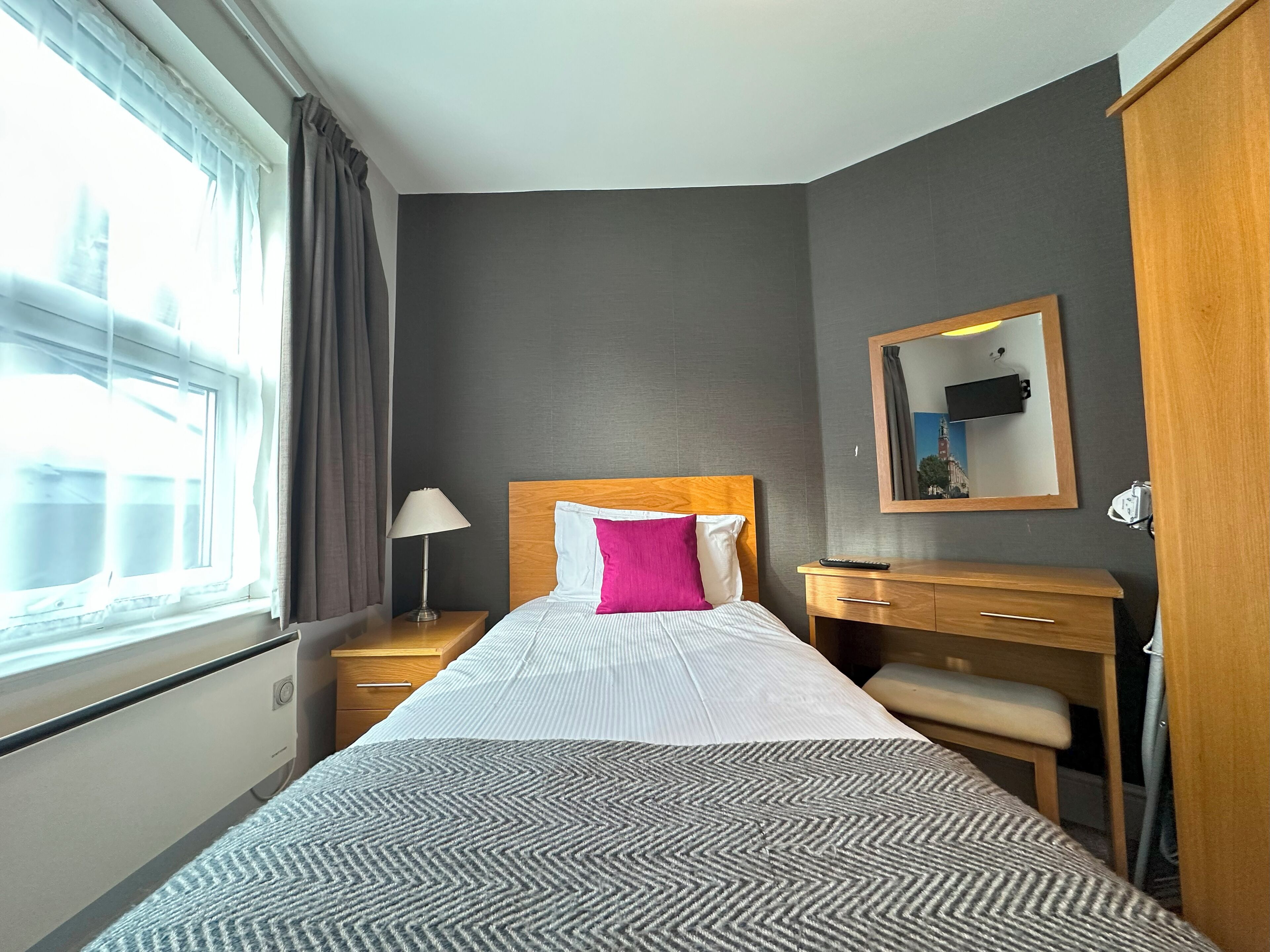Habitación de hotel con cama, mesita de noche, lámpara, espejo y ventana con cortinas.