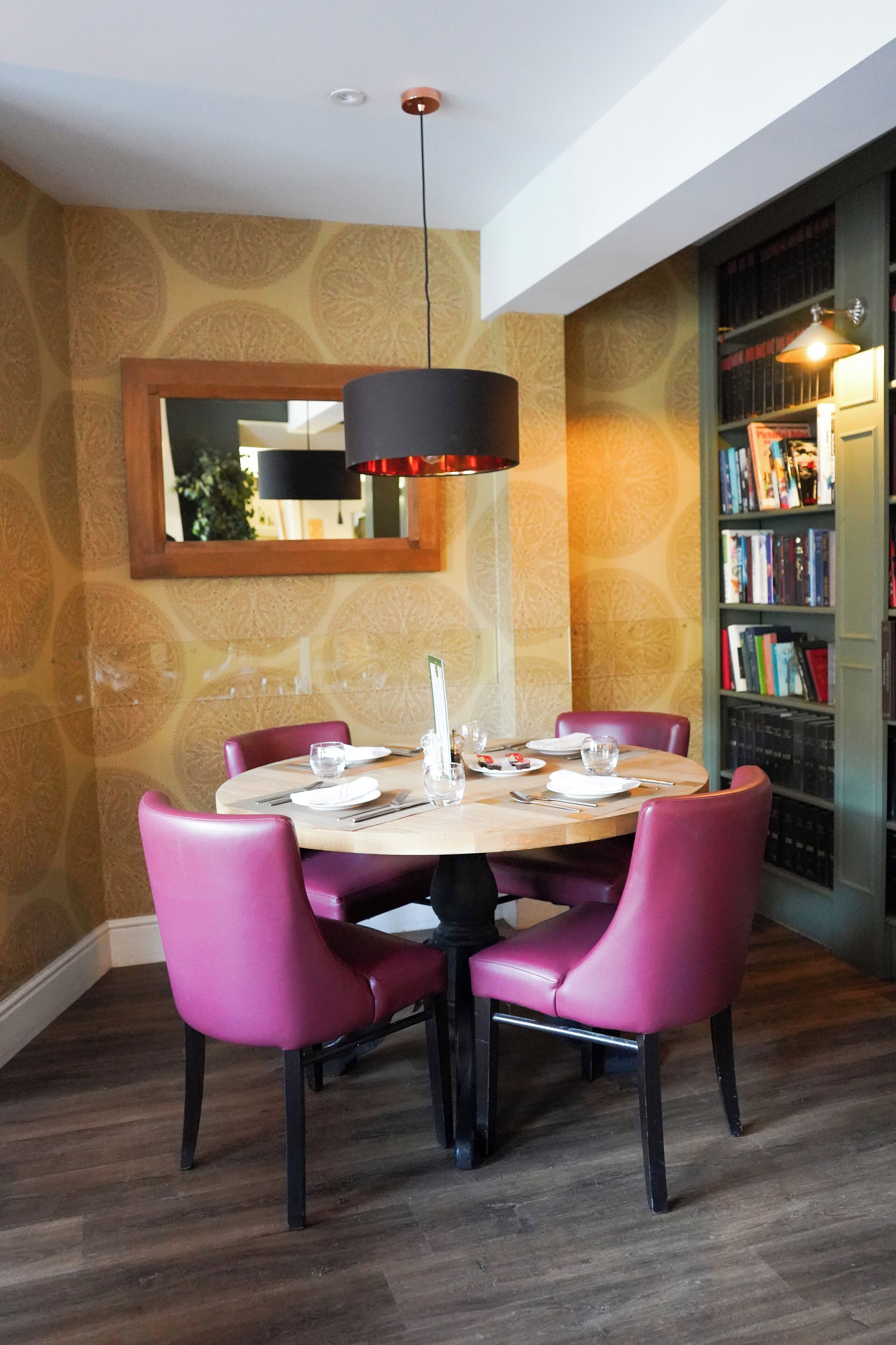 A dining area with a round table, pink chairs, and a bookshelf in the background.