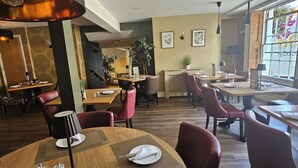 A spacious dining area with round tables and red chairs, featuring a mix of natural and artificial lighting, and a view of the outdoors through large windows. - The North Hill Hotel, Colchester (Colchester)