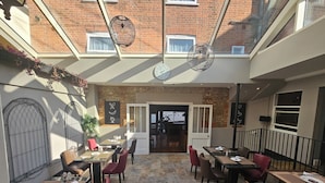 A restaurant with a brick wall, a large clock, and a glass ceiling. Tables and chairs are arranged for dining. - The North Hill Hotel, Colchester (Colchester)