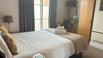 Quarto de hotel com cama, escrivaninha, cadeira, televisão, guarda-roupa e janela com cortinas.