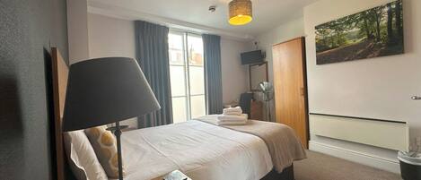 Een hotelkamer met een bed, een bureau met lamp en afstandsbediening, een televisie aan de muur, een schilderij en een raam met gordijnen.