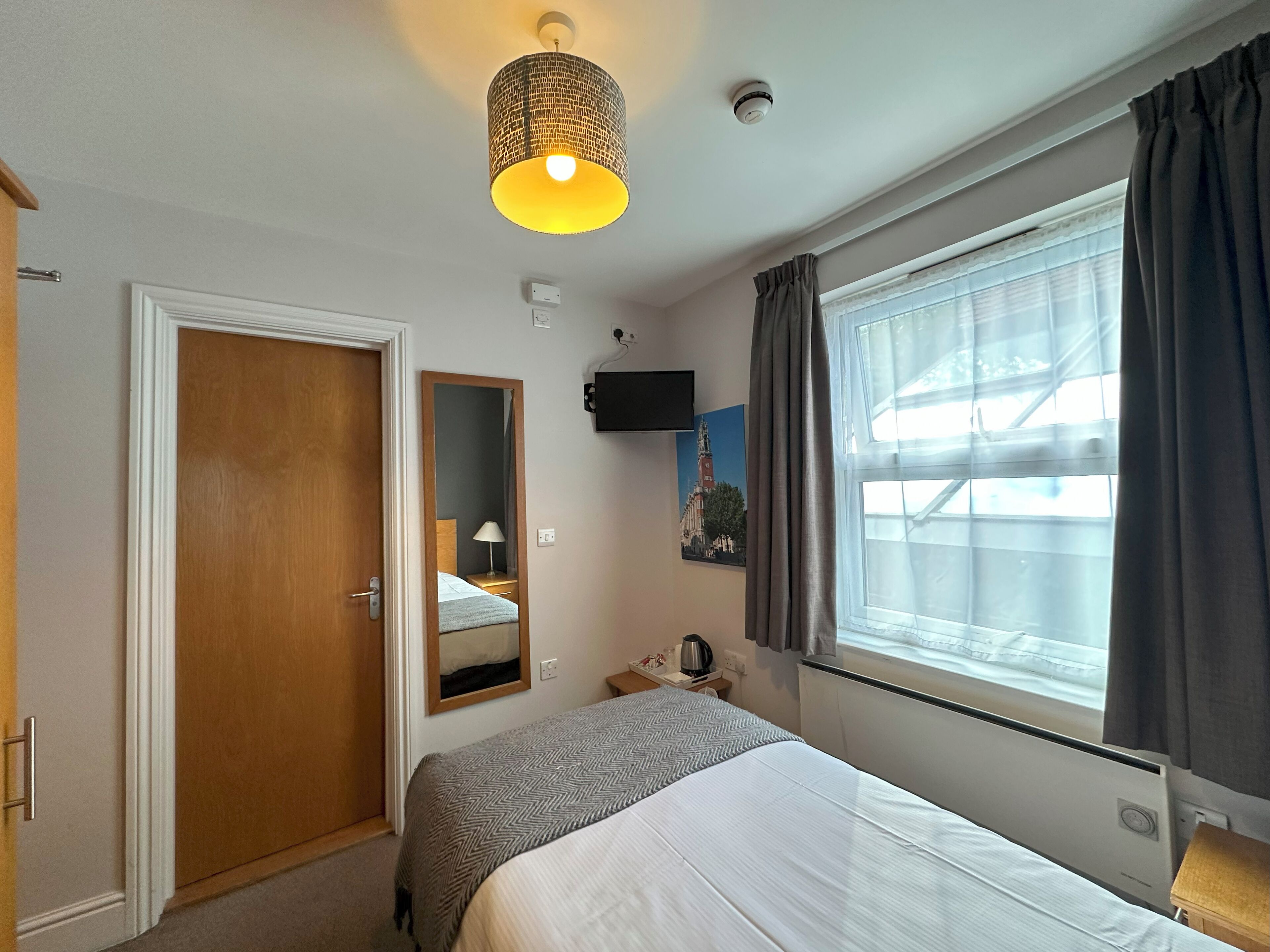 A hotel room with a bed, a bedside table, a lamp, a mirror, a window with curtains, a wall-mounted TV, and a ceiling light fixture.