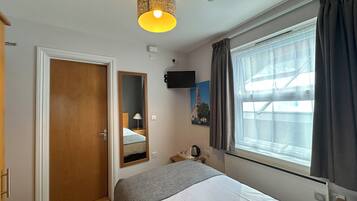 A hotel room with a bed, a bedside table, a lamp, a mirror, a window with curtains, a wall-mounted TV, and a ceiling light fixture.
