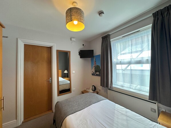 Habitación de hotel con cama, mesita de noche, lámpara, espejo, ventana con cortinas, televisor montado en la pared y lámpara en el techo.