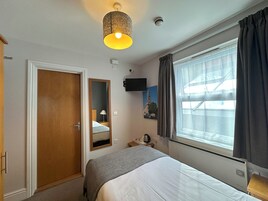Habitación de hotel con cama, mesita de noche, lámpara, espejo, ventana con cortinas, televisor montado en la pared y lámpara en el techo.