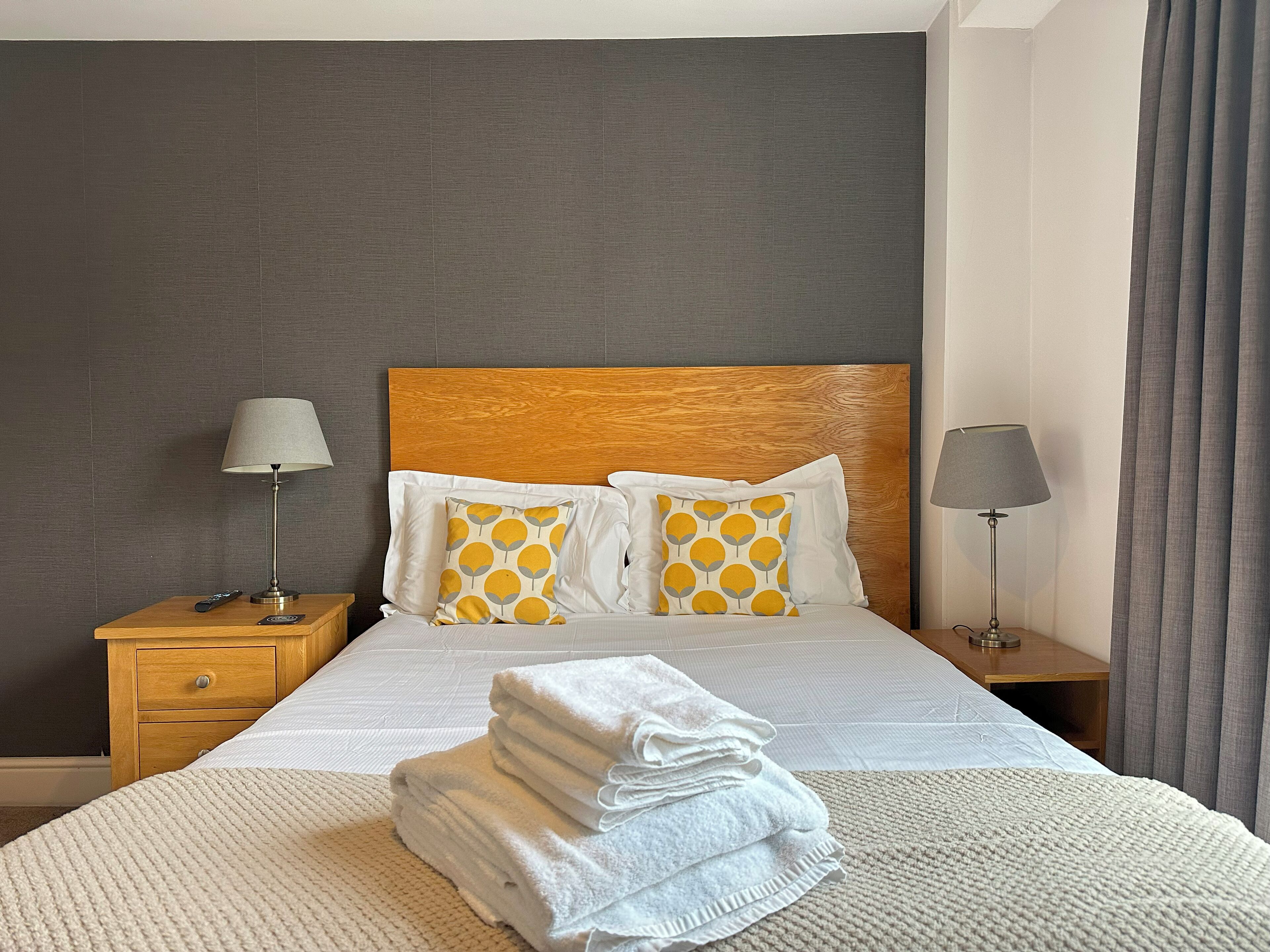 A neatly made bed with white linens and two decorative pillows, a wooden headboard, and a bedside table with a lamp.