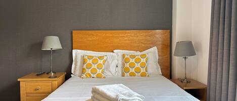 Una cama bien hecha con sábanas blancas y dos almohadones decorativos, cabecera de madera y mesita de noche con lámpara.