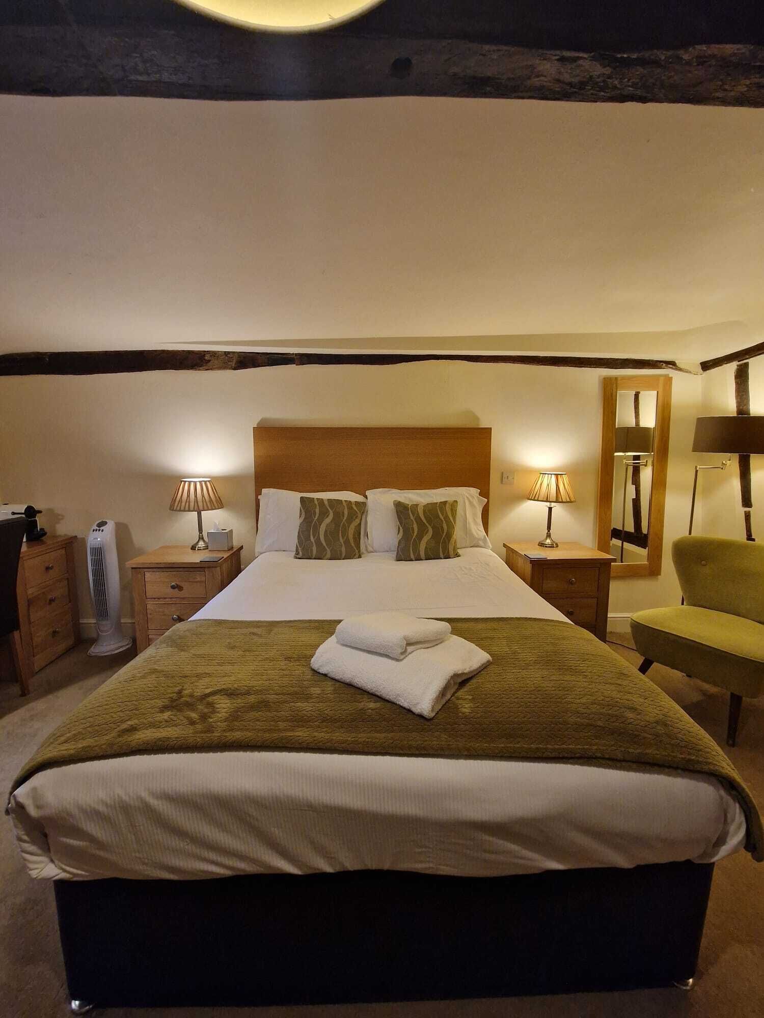 Un lit bien fait, recouvert d’une couverture marron et doté de draps blancs, deux lampes de chevet, une table de chevet en bois, un fauteuil vert et un miroir accroché au mur.