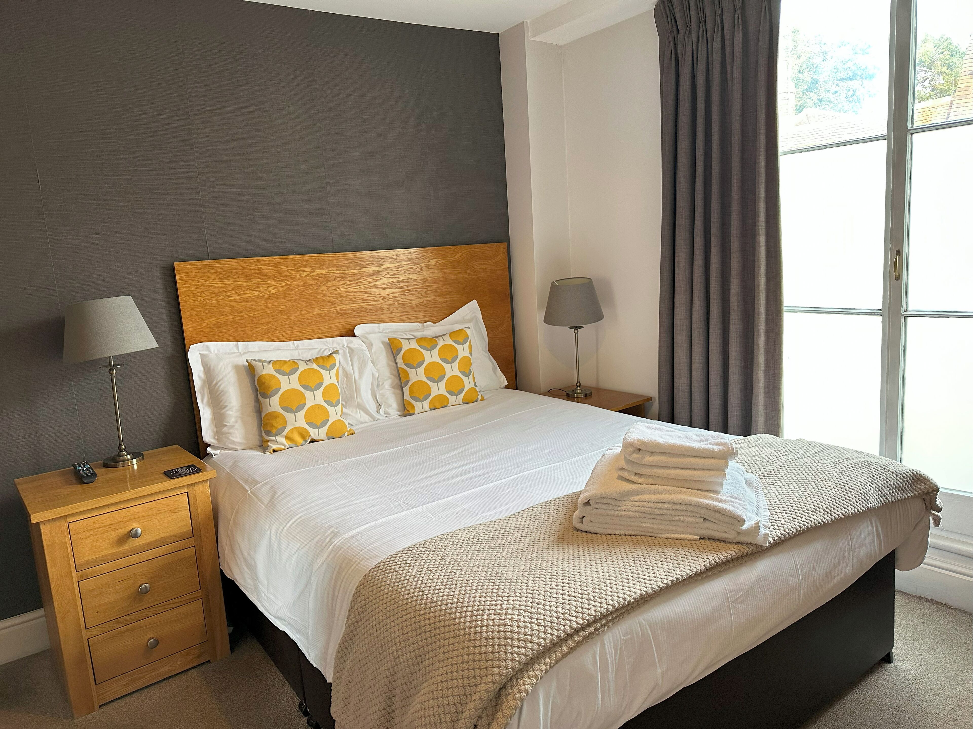 Una cama bien hecha con una manta color beige, dos lámparas de noche y una mesita de noche de madera con cajones.