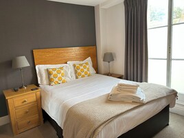 Una cama bien hecha con una manta color beige, dos lámparas de noche y una mesita de noche de madera con cajones.