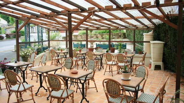 An outdoor patio with wooden tables and wicker chairs, covered by a metal roof, surrounded by potted plants and a stone wall. - Auberge Saint Martin (Saint-Martin-Terressus)