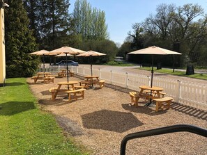 An outdoor seating area with wooden tables and picnic-style benches, surrounded by a white picket fence.