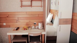 A compact hotel room with a wooden headboard, a small desk with a remote control, a plate of fruit, a kettle, and a bed with white bedding. - Small Hotel Bishkek (Bishkek)