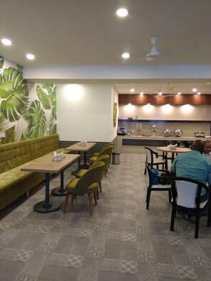 A restaurant with a green sofa, multiple tables, and chairs. There is a kitchen area with a counter and various appliances. - Hotel Palm Greens (New Delhi)