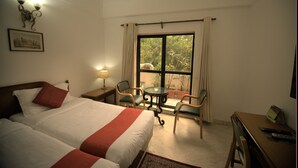 A hotel room with two beds, a small table with chairs, a desk with a chair, a window with curtains, and a lamp. - Hotel Palm Greens (New Delhi)