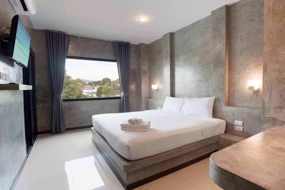 A modern hotel room with a large bed, a TV mounted on the wall, and a view of the city through the window.