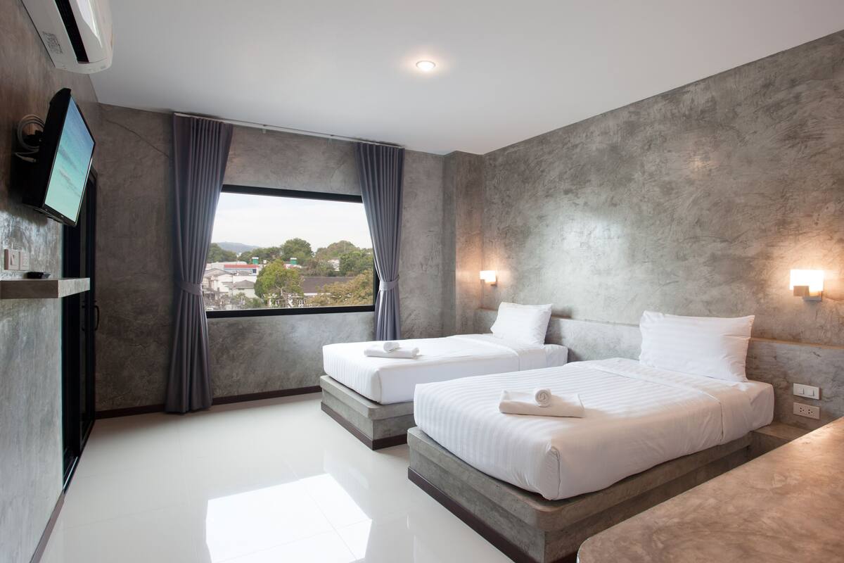 A modern hotel room with two beds, a large window with curtains, a flat-screen TV, and a view of the city.