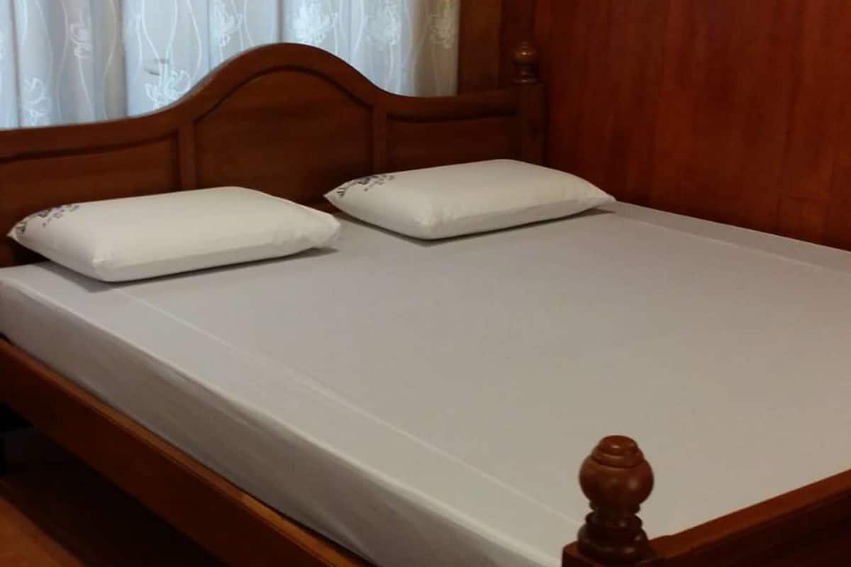 A wooden bed with white bedding and two pillows, set against a wooden wall and a window with patterned curtains.