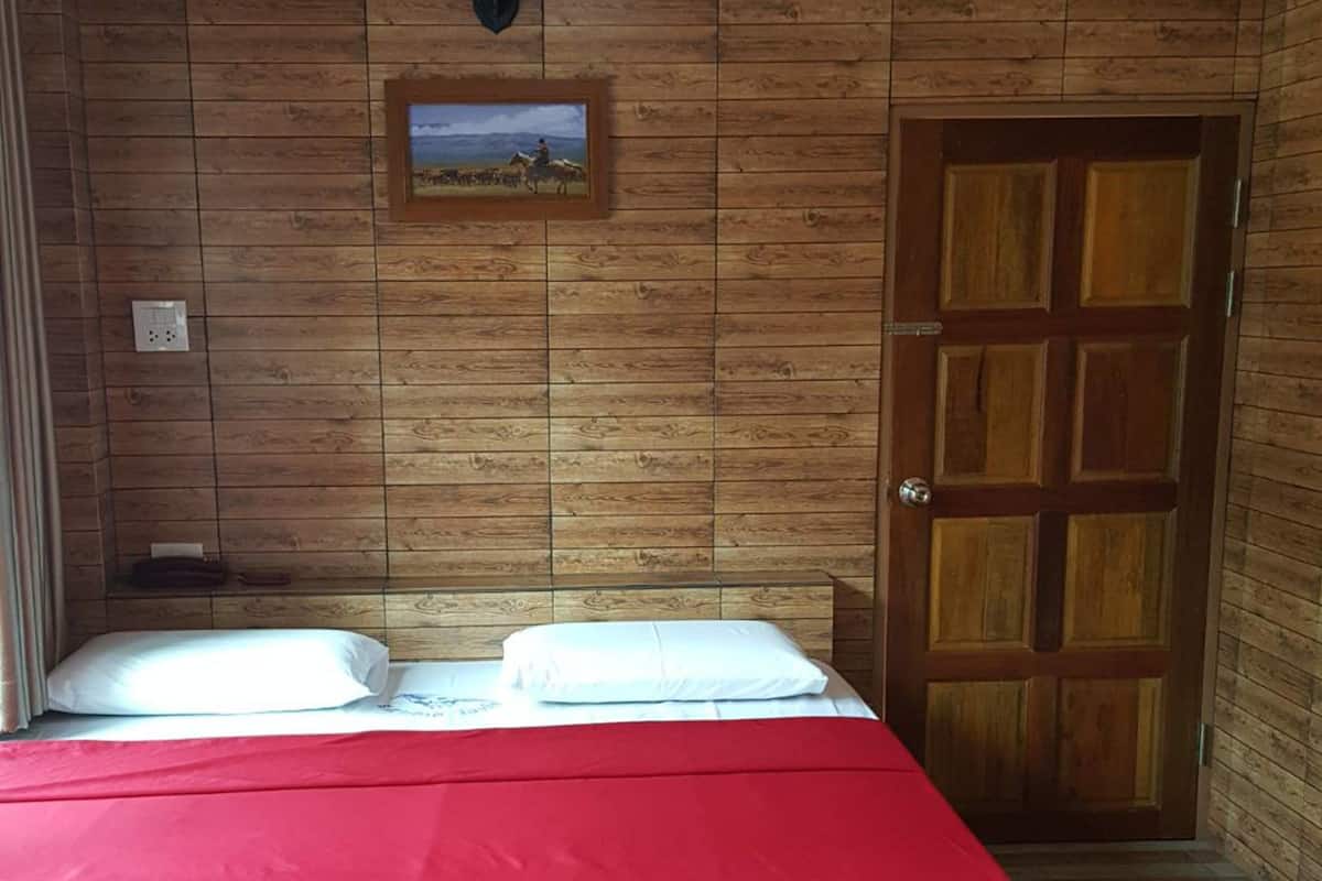 A room with a wooden wall, a bed with a red bedspread, a framed picture, and a wooden door.