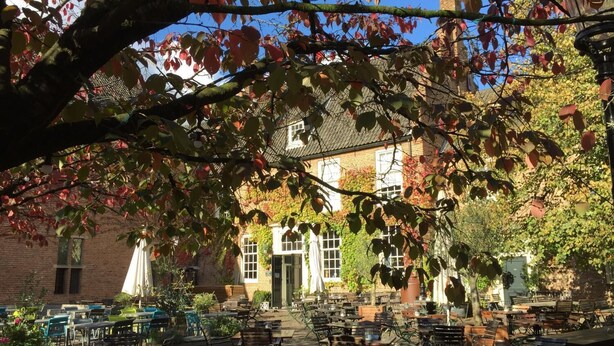 An outdoor café with tables and chairs, a brick building in the background, and trees with autumn leaves.