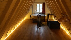A narrow attic room with wooden beams, a window with red curtains, a bathtub, and a small bench.