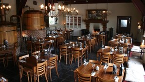 A restaurant with multiple wooden tables and chairs, a brick wall, and a large wooden barrel.