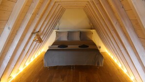 A narrow, wooden-paneled attic bedroom with a bed centered on the floor, flanked by slanted wooden walls and a small window.