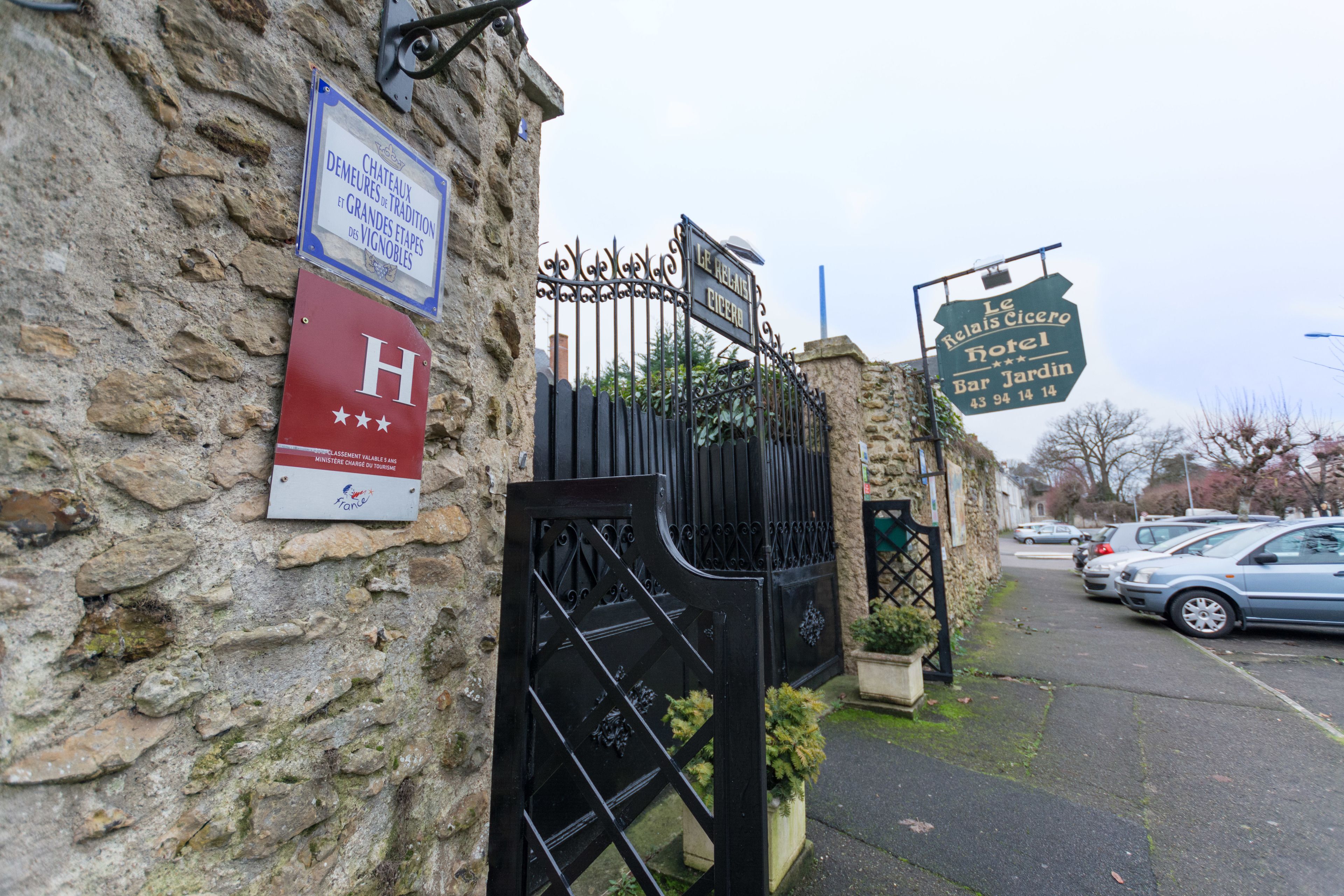 A stone building with a wrought iron gate, a sign for 'Le Relais Cicero Hotel Bar Jardin', and a red sign with a white 'H' and stars.