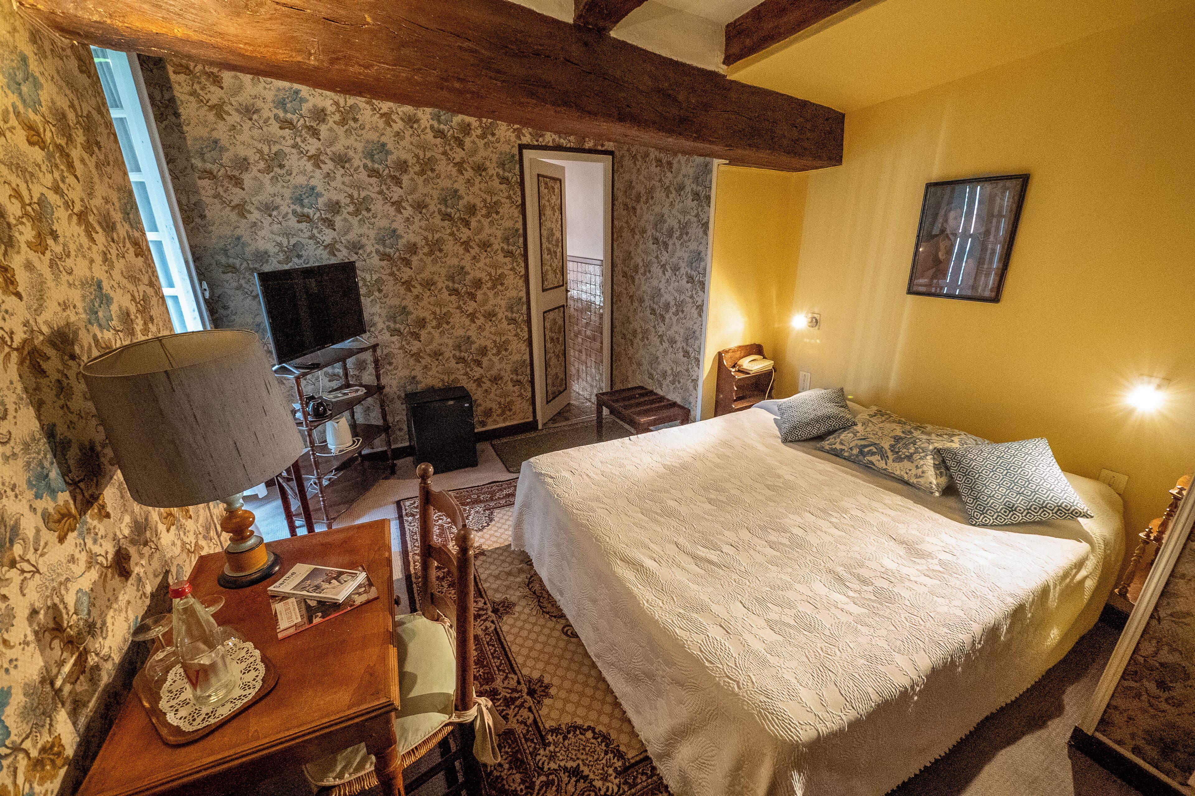 A bedroom with a large bed, a desk with a lamp, a chair, a TV, and a door leading to another room.