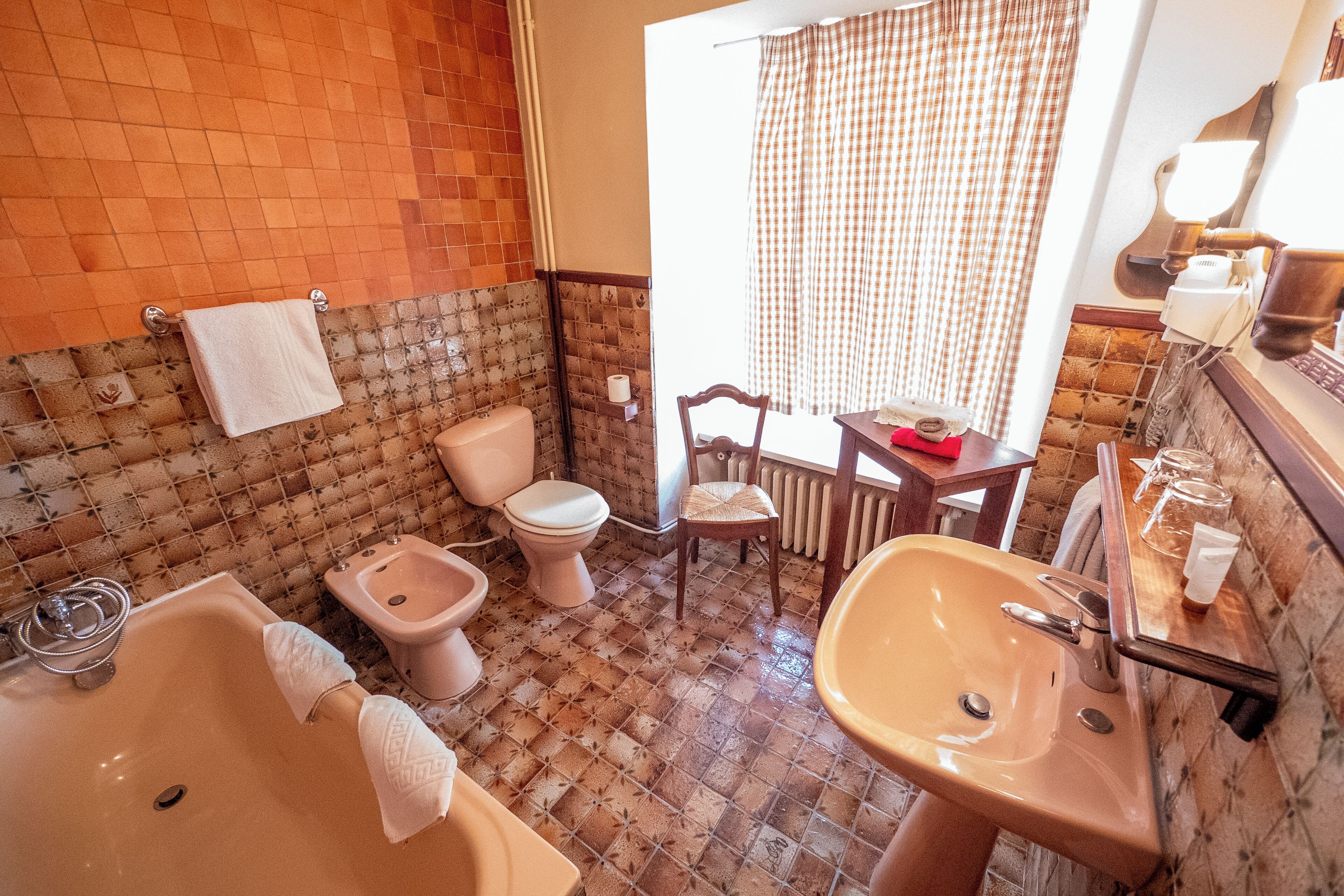 A bathroom with a bathtub, sink, toilet, and bidet. There is a chair and a table with a red object on it.