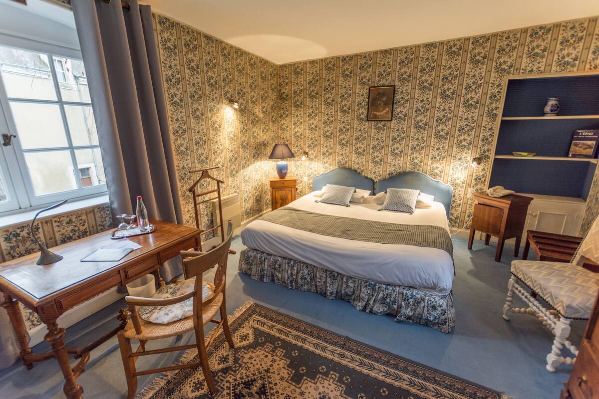 A bedroom with a double bed, a desk with a chair, a bookshelf, and a window with curtains.