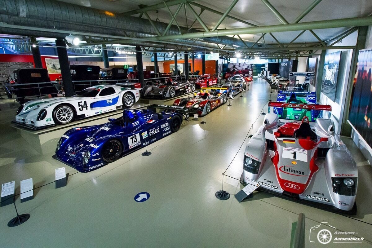 A museum with various racing cars on display, including a white car with the number 54 and a blue car with the number 13.