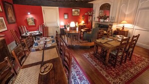 Un coin repas avec une table en bois, dressée pour quatre personnes, entourée de chaises. On y trouve une cheminée avec un manteau et un miroir au-dessus.