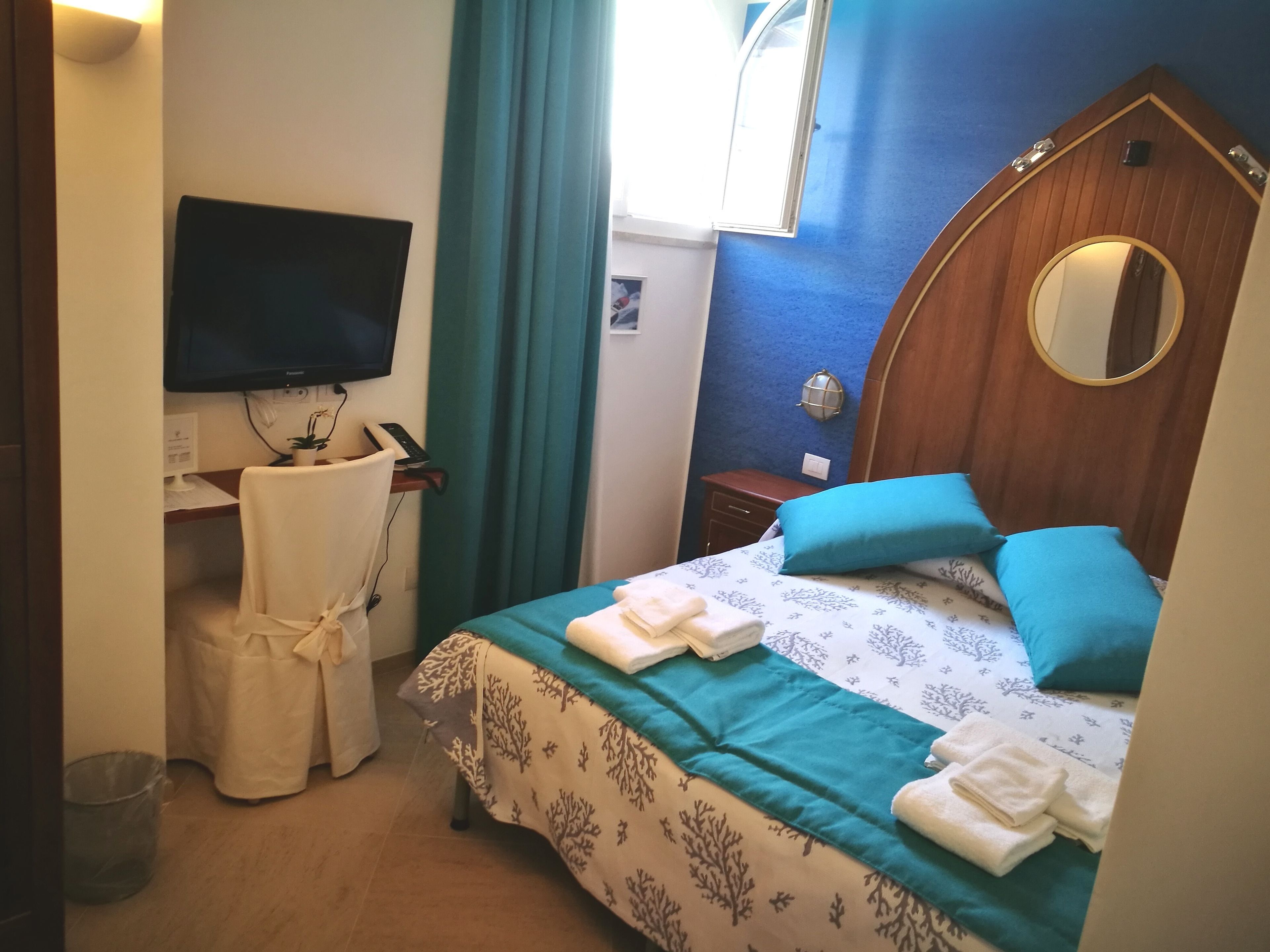 A hotel room with a bed, a desk with a computer, a TV, a mirror, and a window with curtains.