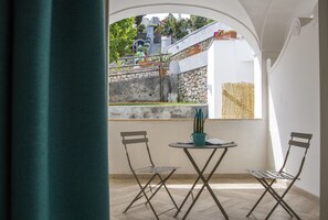 Un pequeño balcón con dos sillas de metal y una mesa, una planta en maceta, y vistas a un jardín con escalones y plantas.