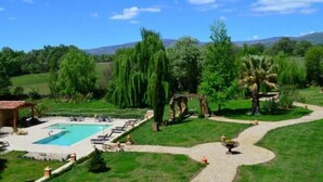 A pool area with a clear blue pool, surrounded by lush greenery and a pathway leading to a gazebo. - La Casona de Valfrío (Cuacos de Yuste)