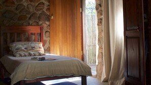 A bedroom with a stone wall, a wooden bed, a thatched roof, and a wooden door.