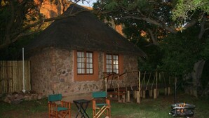 A rustic stone house with a thatched roof, two windows, and a small outdoor seating area with two chairs and a table.