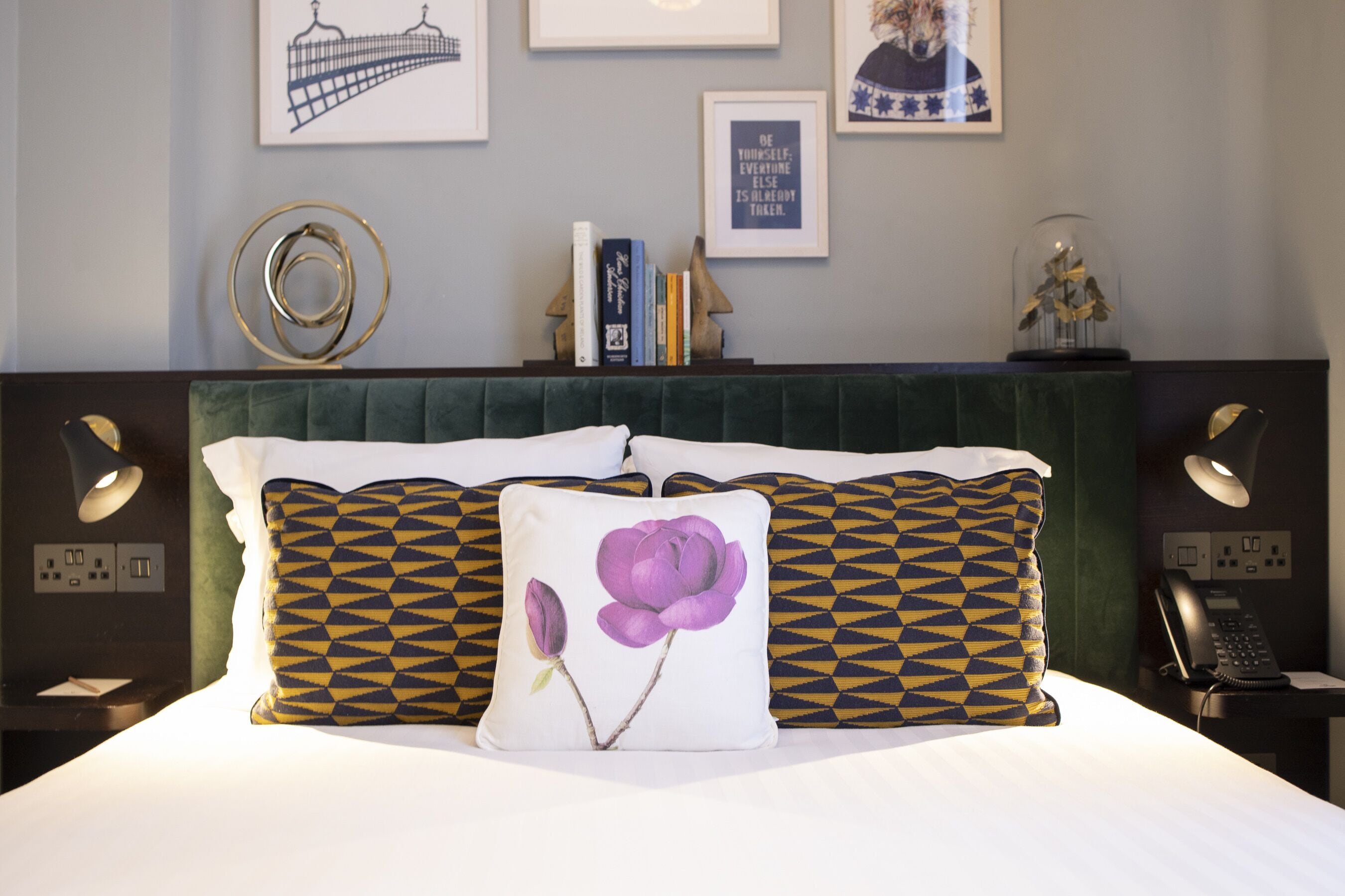 A hotel room with a bed, pillows, a bedside table, a lamp, a phone, and framed pictures on the wall.